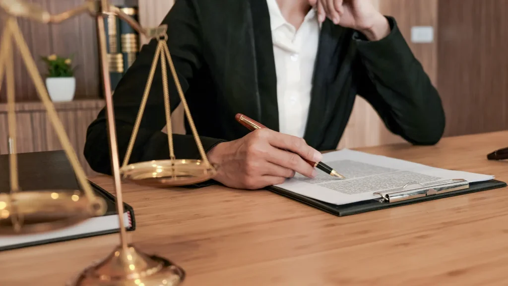 A lawyer in a black suit writing on a document, with a golden scale of justice on the desk.