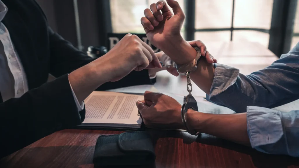 A person in handcuffs is sitting across from another individual, possibly a lawyer or officer, who is discussing a document.