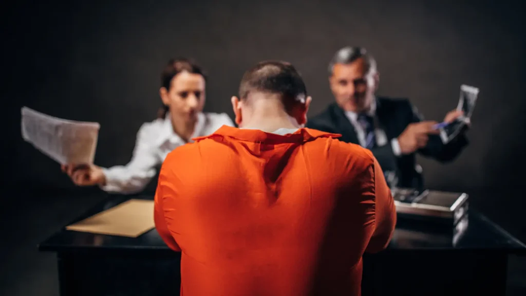 An individual in an orange jumpsuit sits with their back to two people in business attire.